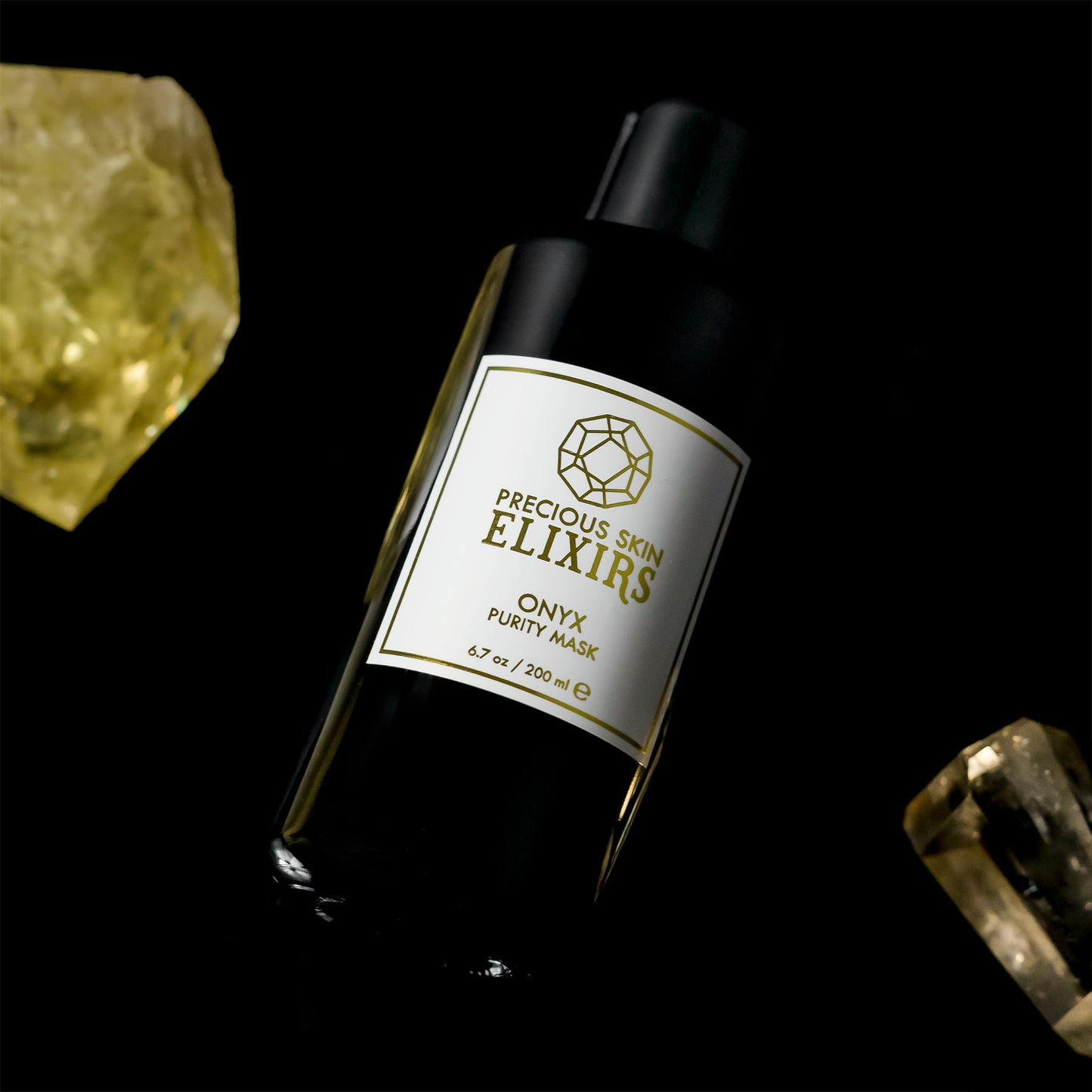Black glass bottle filled with organic face mask powder, chocolate and spaices, rests next to crystals on a black table
